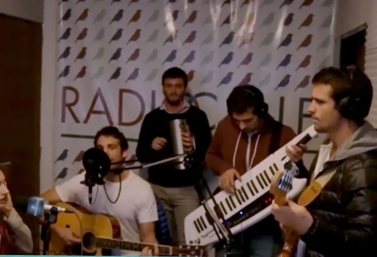 Una banda toca en vivo en el estudio, delante del cartel de Radio a la Calle con pájaros
