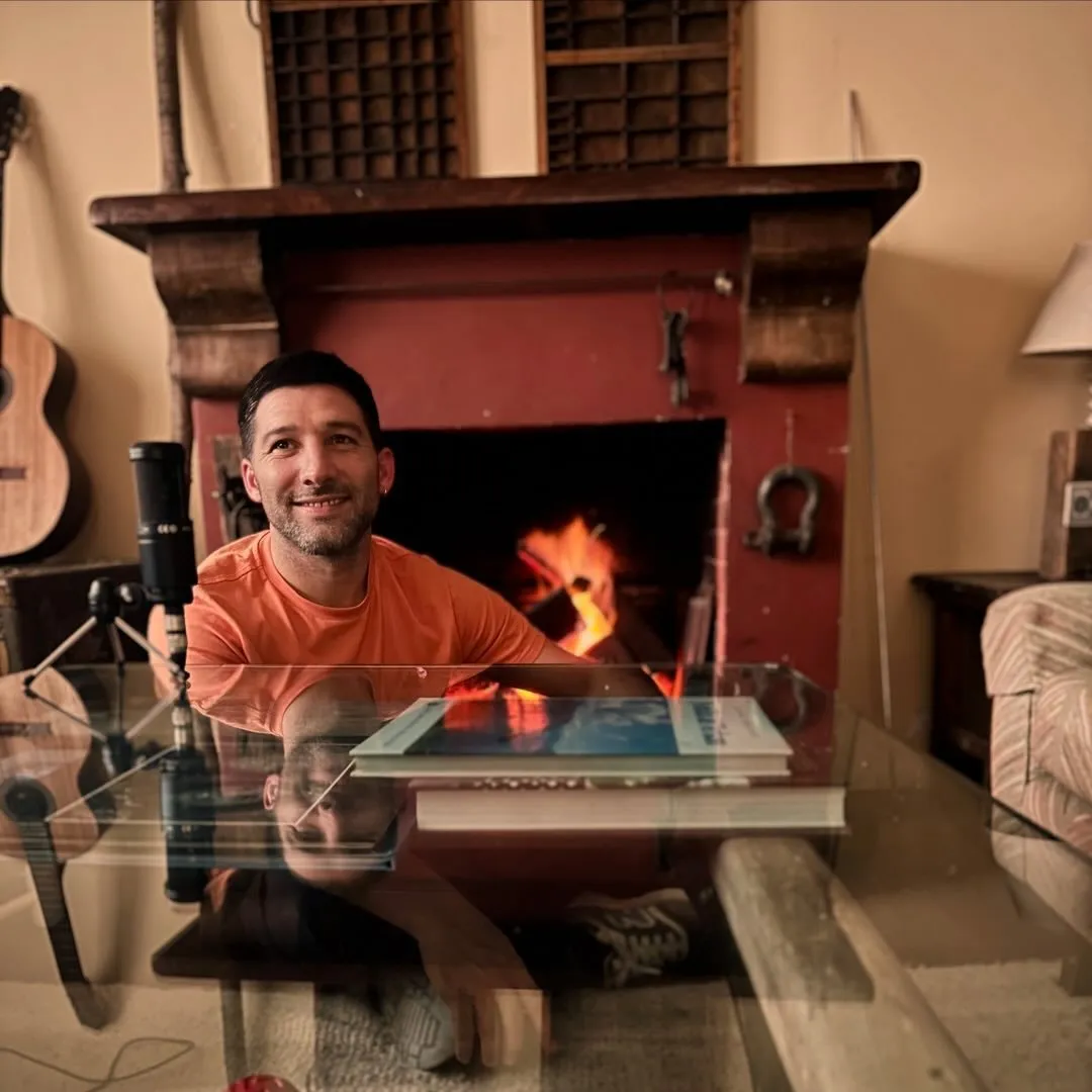Juan Gnarra sonríe frente a una chimenea encendida, con un micrófono y su libro sobre la mesa